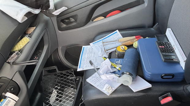 Close-up van de bijrijdersstoel in een werkbus, bezaaid met bonnetjes, gereedschap, pennen, meetlint en rekenmachine.