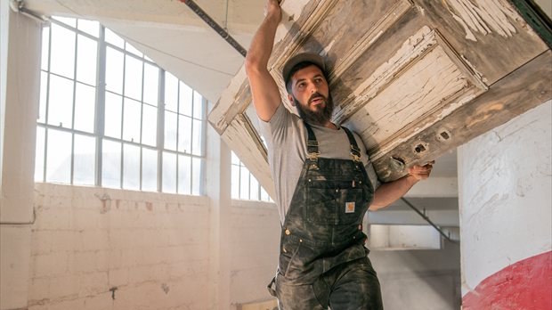 Man draagt oude houten deur op schouder terwijl hij een Carhartt BIB overall draagt