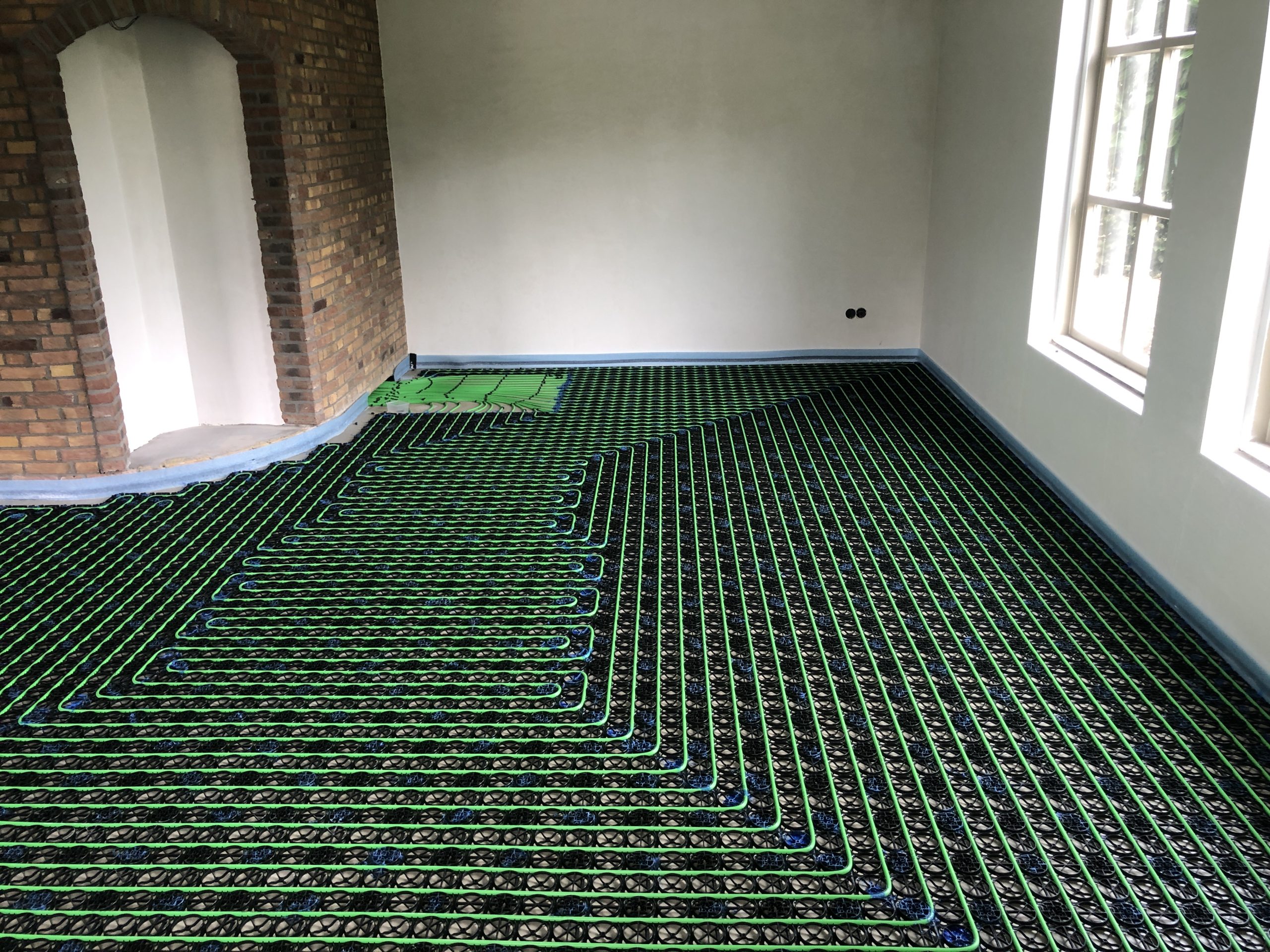 Underfloor radiant heating mesh being installed across a room floor, with green serpentine tubing over a black grid and brick walls nearby.