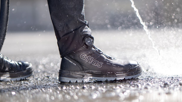 Close-up of a black Atlas athletic shoe stepping on wet pavement with water spraying nearby.