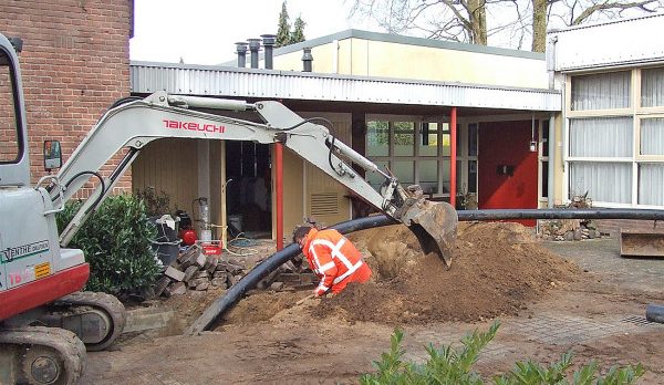 Mini excavator digs a trench beside a brick building, laying a large black pipe while a worker in an orange safety vest guides it.
