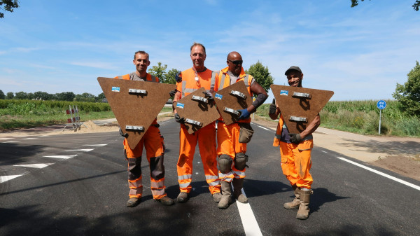Four road workers in bright orange safety gear pose on a freshly paved road, each holding a large wooden shield with metal brackets taken during a road construction project.