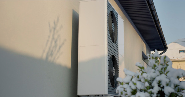 Outdoor air conditioning unit mounted on a building wall with snow-covered shrubs in the foreground.