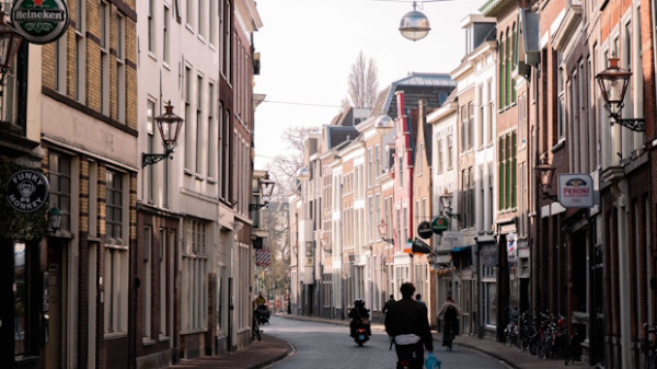 Narrow European street with brick buildings on both sides, pedestrians and cyclists under daylight.
